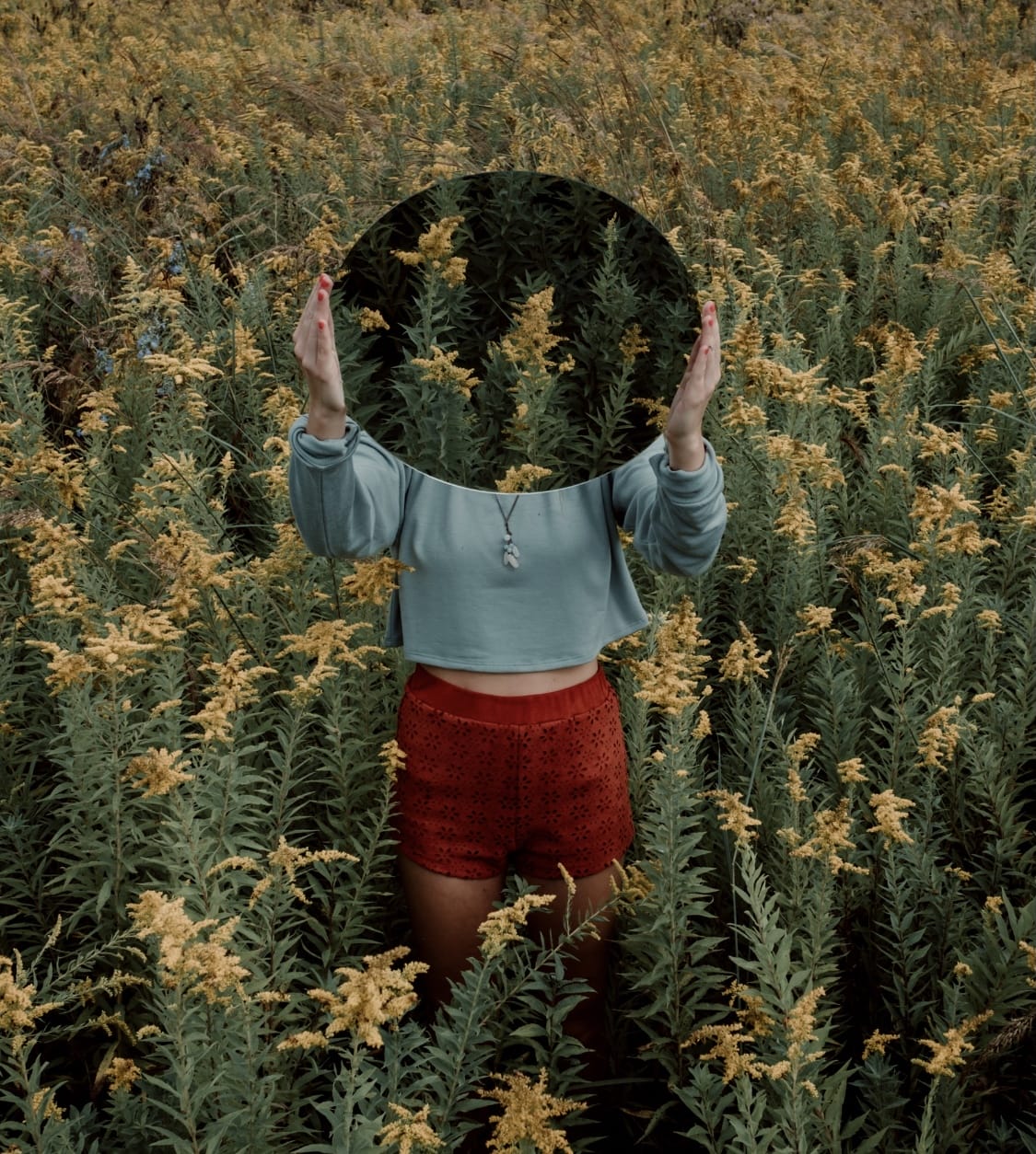 Woman in blue top and red shorts stands in field of yellow flowers with a mirror in front of her face, as if she is searching for her authentic identity