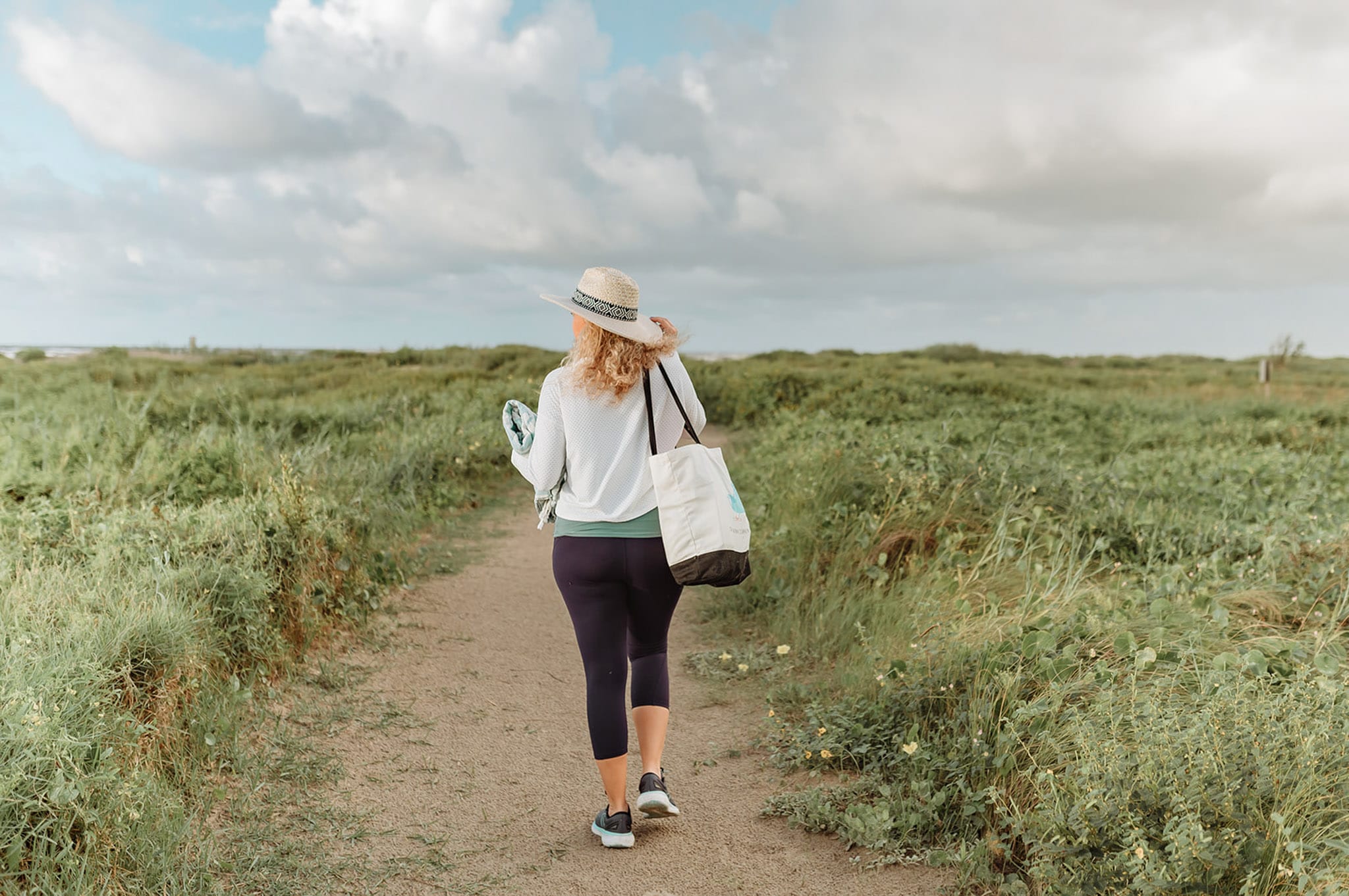 Kim walks down a beach path with a white bag over her shoulder. She's wearing leggings, a white shirt, and a white beach hat.
